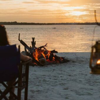 Vrouw die je vanuit haar stoel naar de ondergaande zon kijkt op de Indische Oceaan in Tanzania bij een kampvuurtje