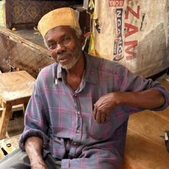 Zittende man met hoedje en hij kijkt je vriendelijk aan bij een kraam op de markt van Stone Town