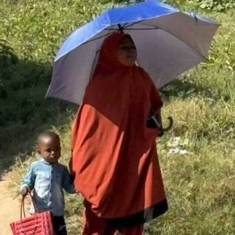 In rood geklede vrouw met paraplu ter bescherming van de zon en aan haar hand een klein jongetje die een tas vasthoudt