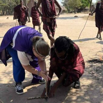 Vrouw die een excursie doet bij de Masai probeert vuur te maken. Een masai helpt haar en daarachter staat een masai toe te kijken