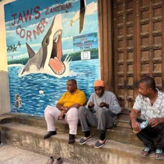 Drie mannen die op een stoepje zitten naast een groot bord waarop staat Jaws corner